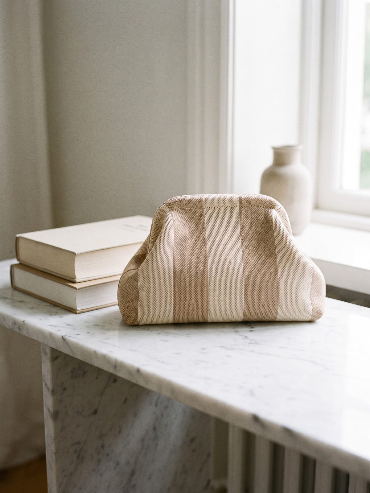 Oat Tonal Stripe Cloud Pouch — sits on a white marble console beside a small stack of two hardcover books of similar height