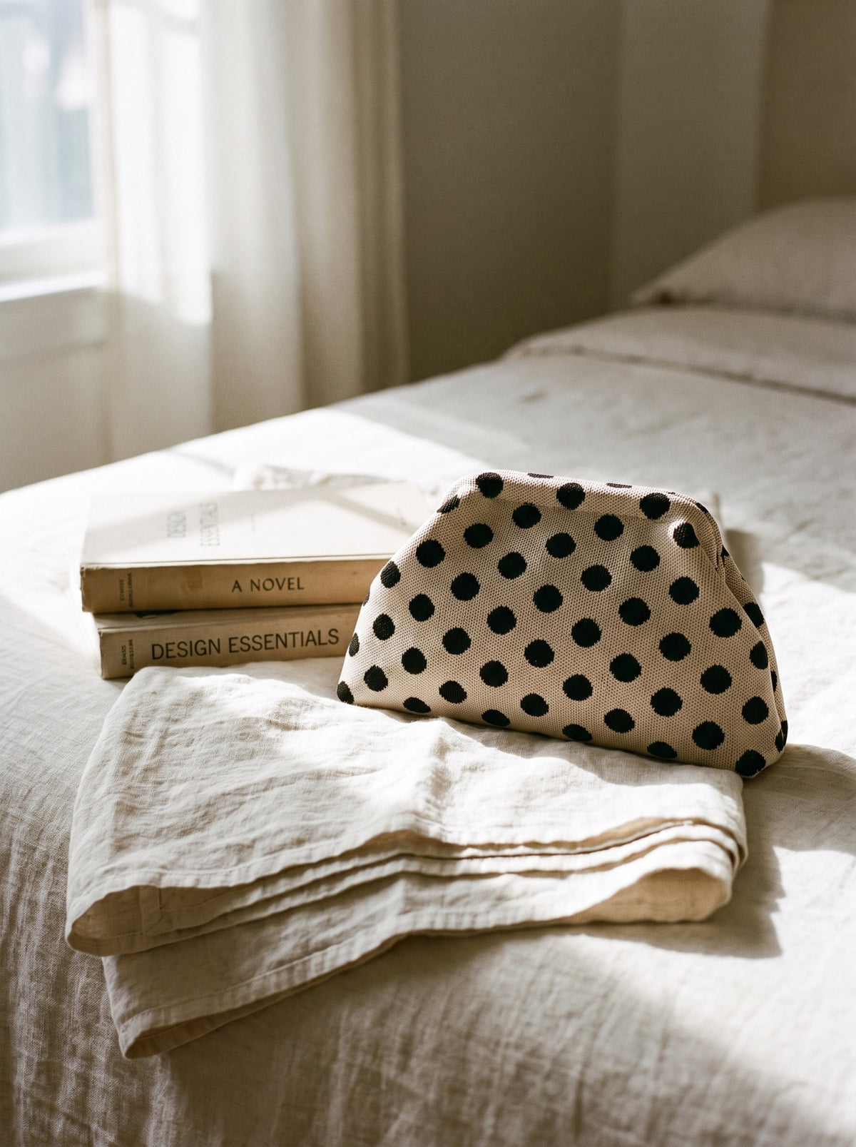 Beige polka dot Etta Rowe cloud pouch resting on a bed beside folded linen and a stack of books
