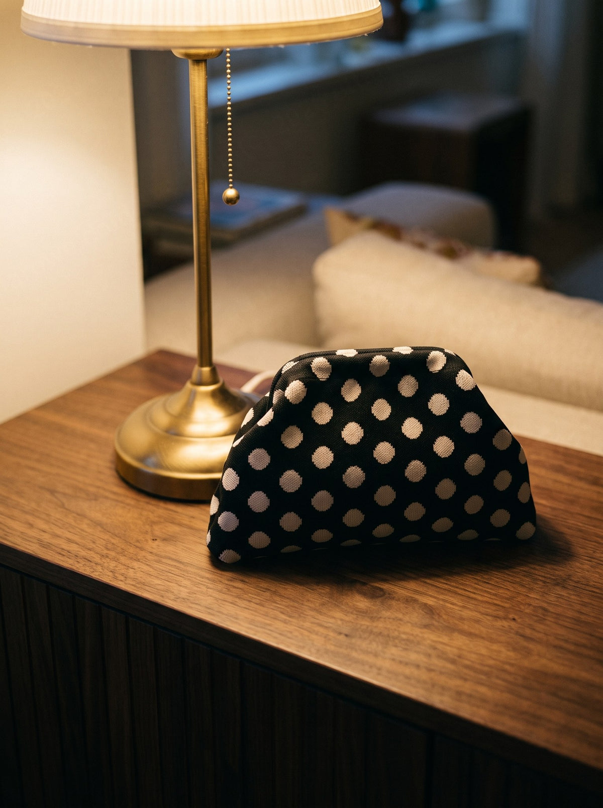 Black polka dot Etta Rowe cloud pouch on a walnut console beside a brass lamp