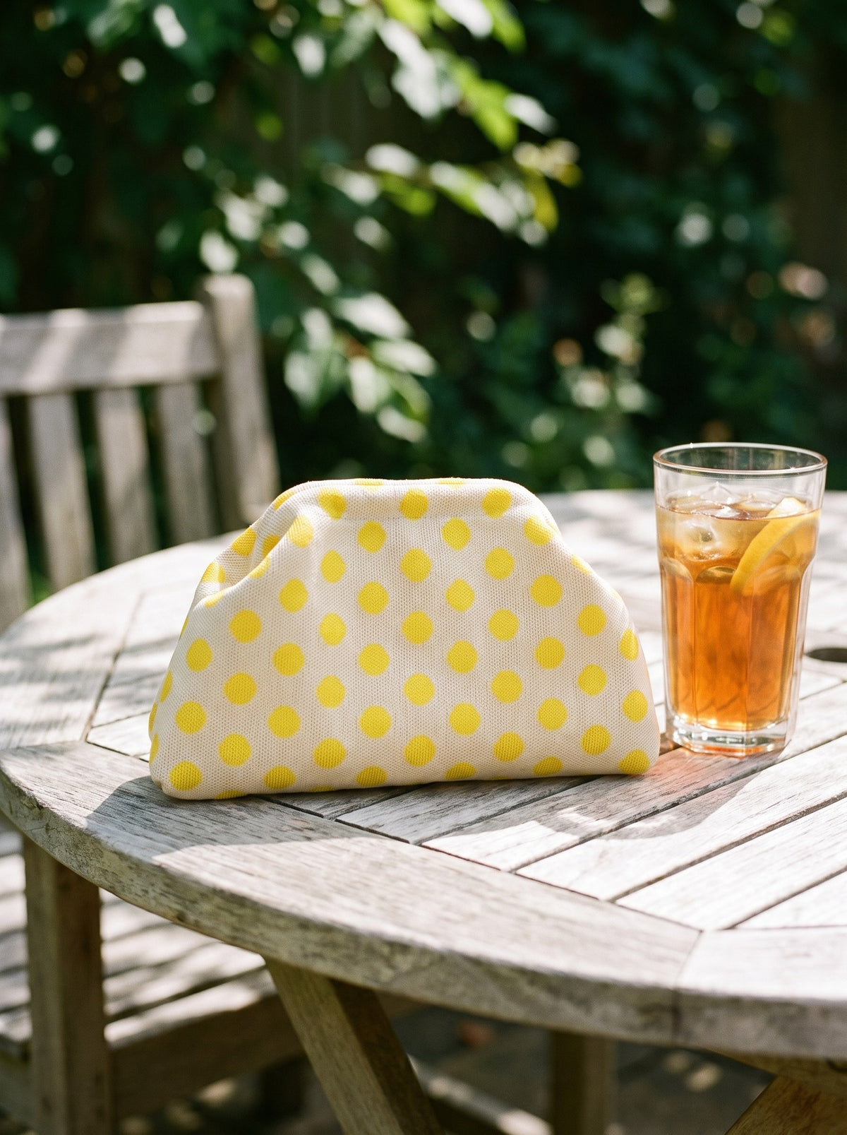 Lemon polka dot Etta Rowe cloud pouch on a garden table beside a glass of iced tea