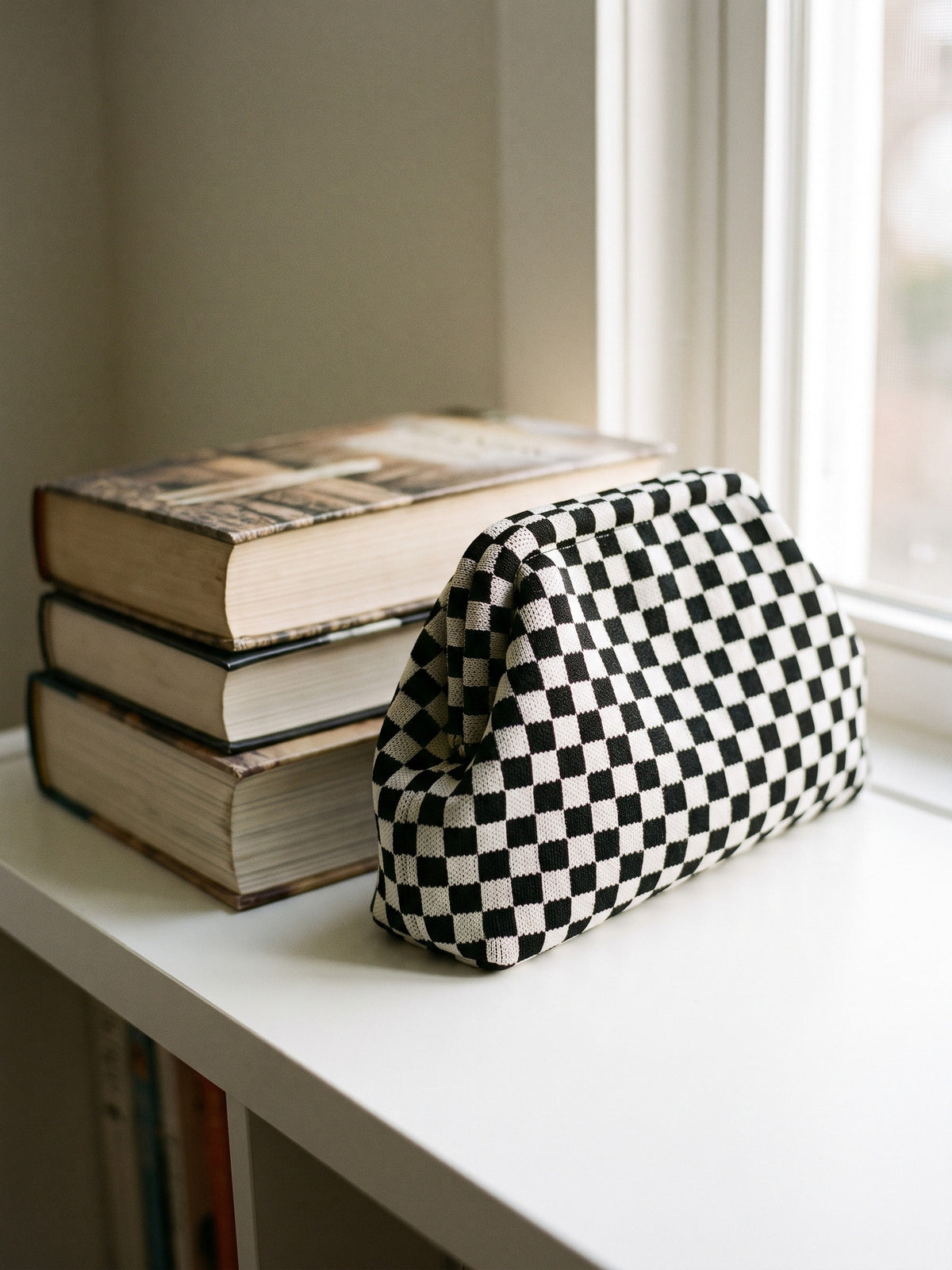 Monochrome Checkerboard Cloud Pouch — stands on a white shelf beside a stack of three hardcover novels