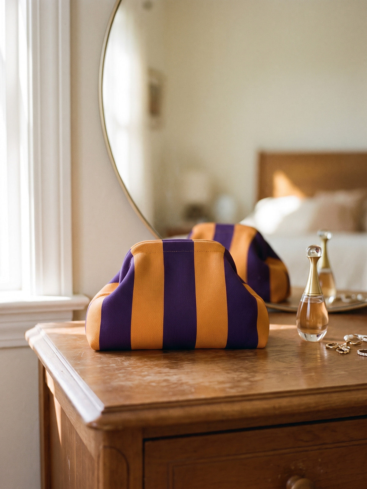 Amber & Plum Stripe Cloud Pouch — stands on a dresser in front of a round mirror, beside a small perfume bottle about half its height