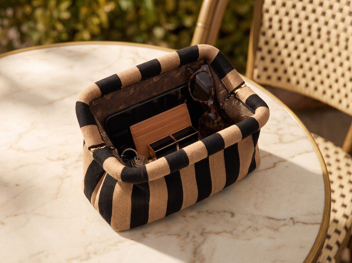 The Camel and Black Stripe Cloud Pouch open on a sunlit marble cafe table, its brass kiss-lock clasp visible, holding a phone, tan cardholder, sunglasses, lipstick and keys